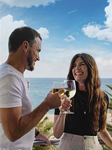 Lachende gasten proosten op een hotelbalkon met uitzicht op zee.